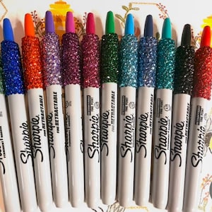 May include: A collection of Sharpie fine point permanent markers with glitter-covered caps in various colors, including green, blue, orange, purple, pink, red, and black. The markers are white with black text and are labeled "RETRACTABLE".