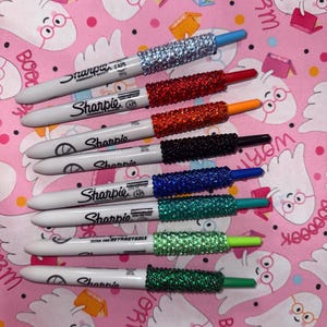 May include: A collection of Sharpie markers with glitter-covered caps in various colors, including silver, red, orange, black, blue, teal, light green, and dark green. The markers are arranged on a pink patterned surface.