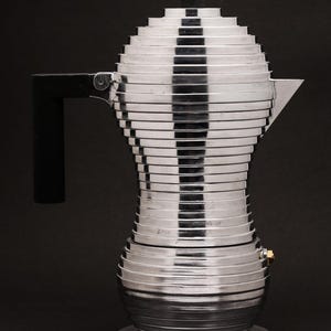May include: A modern, silver-colored espresso maker with a unique, tiered design. It features a black handle and top, and a small gold-colored valve. The coffee maker is made of metal and sits on a round base.
