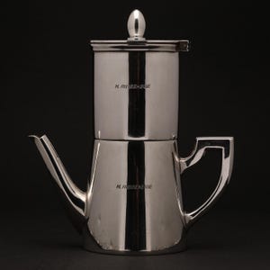 Pode incluir: Uma cafeteira de prata polida com uma secção superior cilíndrica e uma base mais larga e arredondada. Possui um bico, uma pega e uma tampa com um pequeno botão arredondado. O texto "H. RIBBE-EGE" é visível.