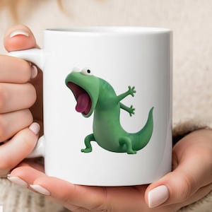 May include: White ceramic mug featuring a cartoon green lizard with a wide open mouth and a pink tongue. The lizard has its arms outstretched. The mug has a handle and the text "MugneticStore" is visible.