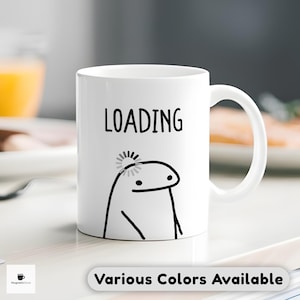 May include: White ceramic mug featuring the word "LOADING" in black, a cartoon character, and a loading symbol. The mug has a curved handle and the text "Various Colors Available" at the bottom.