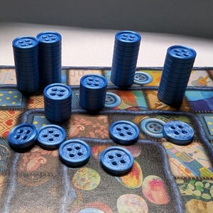 May include: Blue game pieces, resembling buttons, are stacked and scattered on a colorful game board. The buttons have four holes and are various heights. The board has a patchwork design with button illustrations.