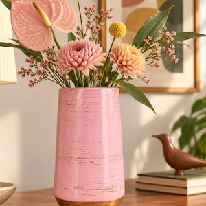 May include: A cylindrical pink vase with a textured, distressed finish and a solid gold-colored base. The vase is filled with a floral arrangement featuring a large pink anthurium, two round yellow billy balls, and two soft pink chrysanthemums.