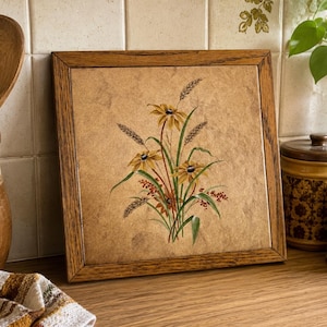 May include: A square decorative tile framed in medium-toned wood featuring a central illustration of yellow coneflowers, green stems, and wheat stalks against a textured, tan, stone-like background. The artwork displays natural botanical details.