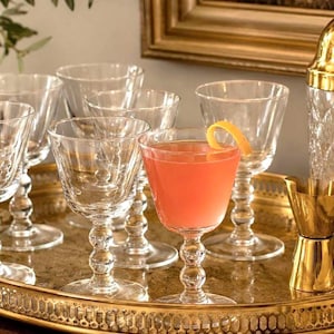 May include: A collection of clear glass stemmed cocktail glasses with a ball-detailed stem, arranged on a gold-toned oval metal tray. One glass is filled with a pink liquid and garnished with a curled orange peel, alongside a gold jigger.