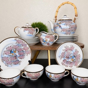 May include: A porcelain tea set featuring a white base with hand-painted blue birds perched on branches with pink blossoms. The set includes a teapot with a bamboo handle, a lidded sugar bowl, a creamer, four teacups, and two matching saucers.