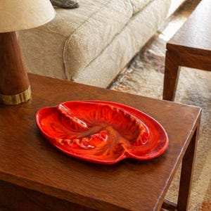 May include: A vibrant red ceramic ashtray with a unique, organic shape, resting on a dark wooden table. The ashtray has a glossy finish and a textured interior, adding visual interest to the mid-century modern design.