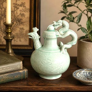 May include: A celadon-colored ceramic teapot with a dragon handle and a bird-shaped spout. The teapot features embossed floral patterns and sits on a wooden surface. A small, decorative plate is visible in the background.