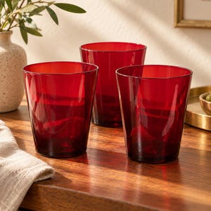May include: Three translucent red glass tumblers with a tapered cylindrical shape and smooth, clear walls. The set consists of two glasses positioned in the foreground and one glass placed slightly behind them, all featuring a simple, open-top design.