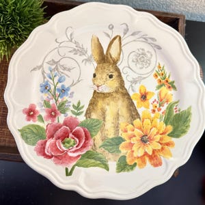 May include: A white ceramic plate with a scalloped edge, featuring a watercolor illustration of a brown bunny surrounded by colorful flowers. The flowers include red roses, yellow daisies, and blue forget-me-nots. Decorative floral accents are also present.