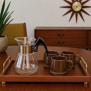 May include: A mid-century modern coffee set on a wooden tray. The set includes a clear glass coffee maker with a black handle, three brown ceramic mugs, and a wooden tray with brass handles. The tray is 12 inches long.