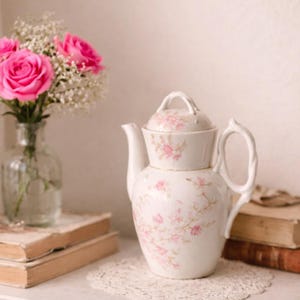 May include: A white ceramic teapot with a lid, adorned with pink floral patterns. The teapot sits on a white lace doily, next to a vase of pink roses and old books. The teapot has a twisted handle.