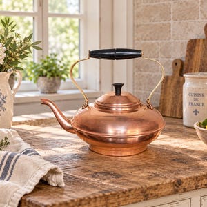 May include: A polished copper tea kettle with a wide, rounded base, a long curved spout, and a black handle. The kettle features a matching black knob on the lid and is supported by a brass-toned metal frame that connects the handle to the body.