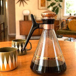 May include: A vintage coffee carafe with a copper lid and accents, a black handle, and a glass body. Two ceramic mugs with geometric patterns sit nearby. The scene is set on a wooden table in a well-lit room.