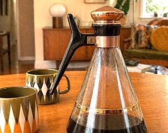 Könnte beinhalten: Eine Vintage-Kaffeekaraffe mit Kupferdeckel und -akzenten, einem schwarzen Griff und einem Glaskörper. Zwei Keramikbecher mit geometrischen Mustern stehen daneben. Die Szene spielt sich auf einem Holztisch in einem hellen Raum ab.