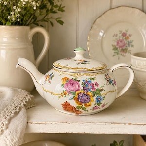 May include: A cream-colored ceramic teapot featuring a colorful floral pattern with pink, yellow, and blue flowers, accented by gold trim along the rim, handle, and spout. The lid has a small green knob and matching floral details.