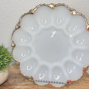 May include: A round white milk glass serving platter featuring twelve oval-shaped indentations arranged in a circular pattern. The platter has a scalloped edge finished with a metallic gold trim, providing a decorative border around the entire piece.
