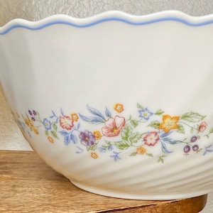May include: White ceramic serving bowl with a wavy blue rim and floral pattern. The bowl features a delicate design of pink, yellow, and purple flowers with green leaves. The bowl has a spiral design on the exterior.