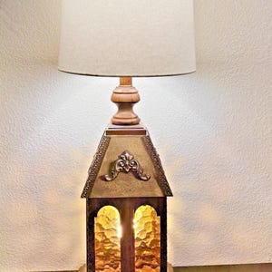 May include: A vintage table lamp with a unique design. The lamp features a beige lampshade, a wooden base, and a decorative metal housing with arched windows. The metal housing has an ornate design and the windows have a textured glass. The lamp is approximately 30 inches tall.