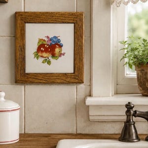 May include: A framed tile featuring a still life of two red apples and a cluster of blue grapes. The artwork is set against a white background and framed in a rustic, light brown wooden frame. The tile is mounted on a white tiled wall.