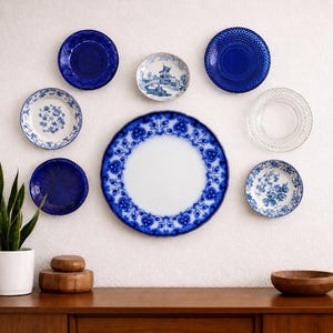 May include: A collection of decorative plates in various sizes and designs, mounted on a white wall. The plates feature blue and white patterns, including floral motifs, textured surfaces, and a central plate with a detailed border. A wooden cabinet and plant are in the foreground.