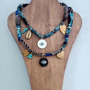 Puede incluir: Dos collares en capas con cordones de tela azul, negra y blanca. Los collares presentan dijes dorados, incluyendo un corazón, un trébol de cuatro hojas, una concha y un colgante en forma de ojo. Los collares se exhiben en un soporte de madera.
