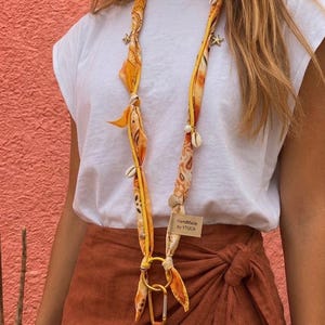 Peut inclure: Un cordon en tissu jaune à motif cachemire orné de petites breloques en forme d'étoile de mer métalliques et de coquillages blancs. Le cordon comprend une étiquette rectangulaire avec le texte HandMade by YTUCA et un anneau doré.