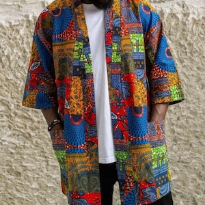 Puede incluir: Una chaqueta estilo kimono abierta por delante con un vibrante patrón de patchwork en tonos rojos, azules, amarillos y verdes. La prenda tiene mangas tres cuartos, dos grandes bolsillos delanteros y se lleva sobre una camiseta blanca.