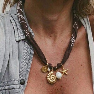 Puede incluir: Un collar de bandana marrón con colgantes dorados, que incluyen una concha, una estrella de mar y un disco con la letra "D". El collar también presenta una perla blanca. La bandana está envuelta alrededor del cuello.