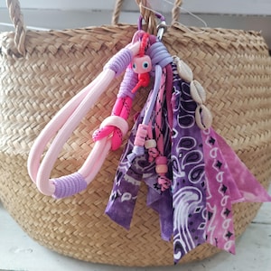 Puede incluir: Cesta de paja tejida con un adorno decorativo. El adorno presenta cuerdas, telas y cuentas rosas y moradas, junto con un pequeño adorno caprichoso en forma de ojo. El adorno también incluye conchas marinas y tela con estampado de cachemira.