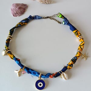 Puede incluir: Un collar hecho a mano con una banda de tela con estampados azules, amarillos y naranjas. El collar presenta dijes dorados, incluyendo una concha, una estrella de mar y un colgante con un diseño de ojo turco. Una pieza de joyería única.