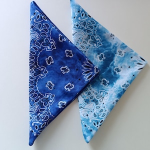 Può includere: Due fazzoletti bandana blu, piegati a triangolo. Uno è blu scuro con un motivo paisley bianco, l'altro è blu chiaro con effetto tie-dye e dettagli bianchi. Ideali per vari usi.