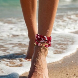 Peut inclure: Bracelets de cheville avec un tissu à motifs rouge et blanc, des perles roses et un charm en forme de croissant de lune doré. Un charm en forme d'étoile dorée est également visible. Les bracelets sont portés sur une plage de sable près de l'océan.