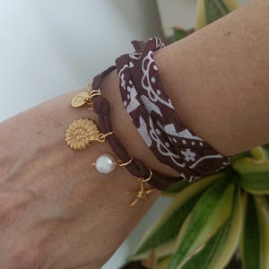 Könnte beinhalten: Armbänder mit braunen Stoffwickeln und goldenen Anhängern, darunter eine Muschel, eine Perle und ein Seestern. Ein Armband ist ein braunes und weiß gemustertes Bandana.