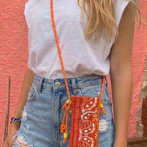 May include: A white t-shirt and light blue distressed denim shorts paired with an orange paisley print crossbody phone pouch. The pouch features a long orange rope strap with yellow and orange beads and a small silver star charm attached.