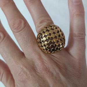 Puede incluir: Un anillo grande, dorado, con un diseño abovedado texturizado. El anillo presenta un patrón repetitivo de pequeñas formas cuadradas, creando una superficie texturizada. El anillo se lleva en un dedo.