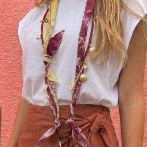 Può includere: Un lungo cordino in tessuto con motivo tie-dye giallo e bordeaux, decorato con piccoli ciondoli dorati tra cui conchiglie e stelle. Il tessuto è annodato in basso a un anello circolare nero collegato a una custodia per telefono rossa.