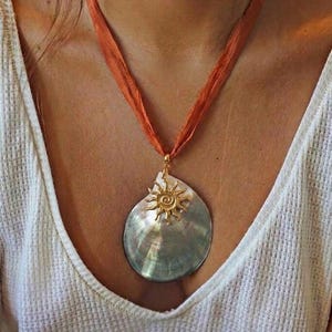Op de afbeelding: Een hangerketting met een grote, iriserende schelpvormige bedel. Een goudkleurig zonontwerp siert de schelp, die aan een oranje lint is bevestigd. De ketting is een opvallend sieraad, perfect voor een vleugje bohemien.