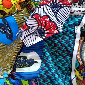 May include: A selection of vibrant African wax print fabrics. The textiles display diverse patterns, such as floral, geometric, and bird motifs. Colours include red, blue, yellow, and green. Some fabrics have printed text.