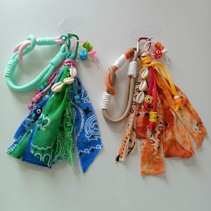 Puede incluir: Dos llaveros hechos a mano con borlas de tela coloridas, conchas marinas y cuentas. Uno presenta un diseño de cuerda verde y rosa, mientras que el otro tiene un diseño de cuerda naranja y beige. Ambos tienen un cierre de metal.