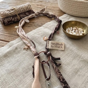 Può includere: Un cordino realizzato con strisce di tessuto intrecciate beige e marrone scuro, decorato con piccoli ciondoli a forma di conchiglia e stella marina. Un anello in metallo marrone unisce il tessuto a un moschettone e una clip trasparente. L'etichetta riporta HandMade by YTUCA.