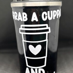 Könnte beinhalten: Schwarzer Reise-Kaffeebecher mit schwarzem Deckel und silbernem Rand. Der Becher zeigt eine weiße Grafik einer Kaffeetasse mit einem Herz und den Worten "GRAB A CUPPA AND..."