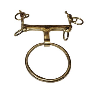 May include: A gold-coloured metal horse bit featuring a large circular ring at the bottom and smaller rings on each side. The bit has a horizontal bar with curved ends and a central vertical bar.