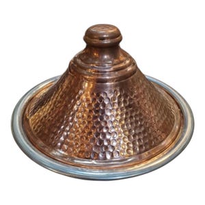 May include: A copper-colored tagine pot with a hammered texture and a conical lid. The pot has a silver-colored rim and a rounded top. This traditional cookware is used for slow-cooked stews and tagines.