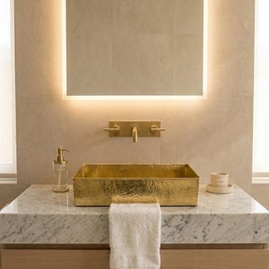 Peut inclure: Une vasque rectangulaire en laiton martelé repose sur un comptoir en marbre blanc avec un meuble-lavabo en bois clair. Au-dessus, un miroir carré rétroéclairé par LED est fixé sur un mur carrelé beige, avec un robinet mural en laiton.