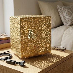Puede incluir: Una lámpara dorada en forma de cubo con intrincados patrones calados, colocada sobre una mesita de noche de madera. El cable y el enchufe de la lámpara son visibles. Libros, un cuenco pequeño y gafas también están sobre la mesa. La lámpara proyecta sombras decorativas.