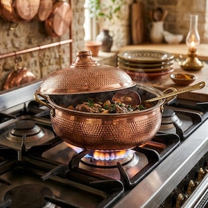 Peut inclure: Une marmite en cuivre martelé avec un couvercle bombé assorti est posée sur un brûleur de cuisinière à gaz. La marmite possède deux poignées latérales et contient un plat cuisiné avec une cuillère de service en métal à l'intérieur.
