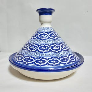 Könnte beinhalten: Ein weißer Keramik-Tajine-Topf mit einem konischen Deckel mit blau-weißen Mustern und einem breiten Boden. Der Deckel hat eine abgerundete Oberseite und einen weiß-blauen Hals. Der Topf hat komplizierte blaue geometrische Designs.