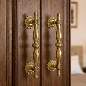 Puede incluir: Primer plano de dos manijas de puerta doradas y ornamentadas sobre una puerta de madera marrón oscuro. Las manijas tienen un diseño vintage detallado con detalles florales y una forma vertical y alargada. La puerta parece ser parte de un armario.